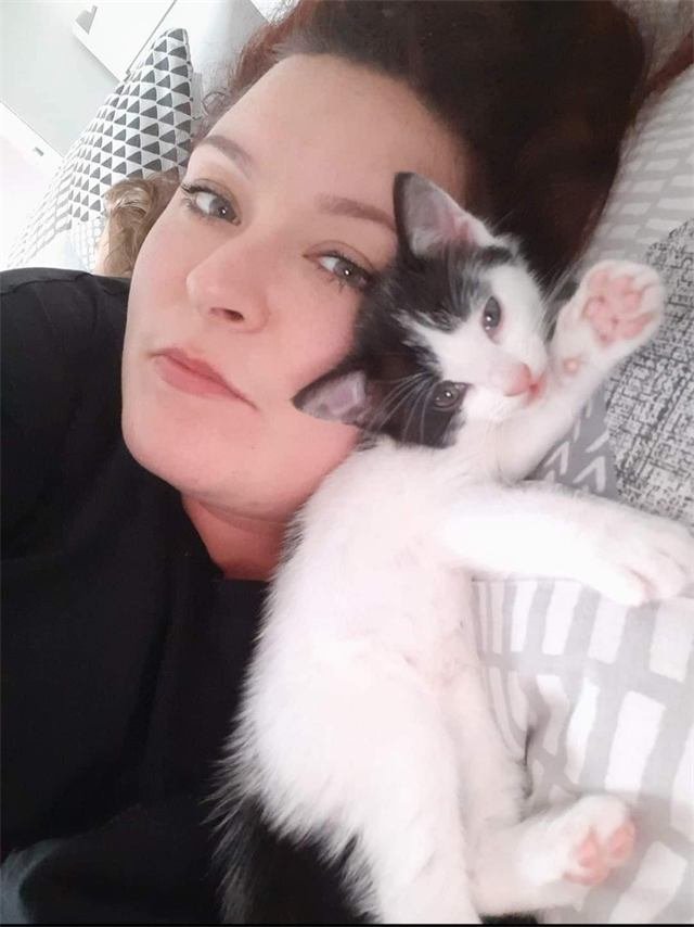 A black and white kitten snuggled against a woman's cheek with one paw in the air